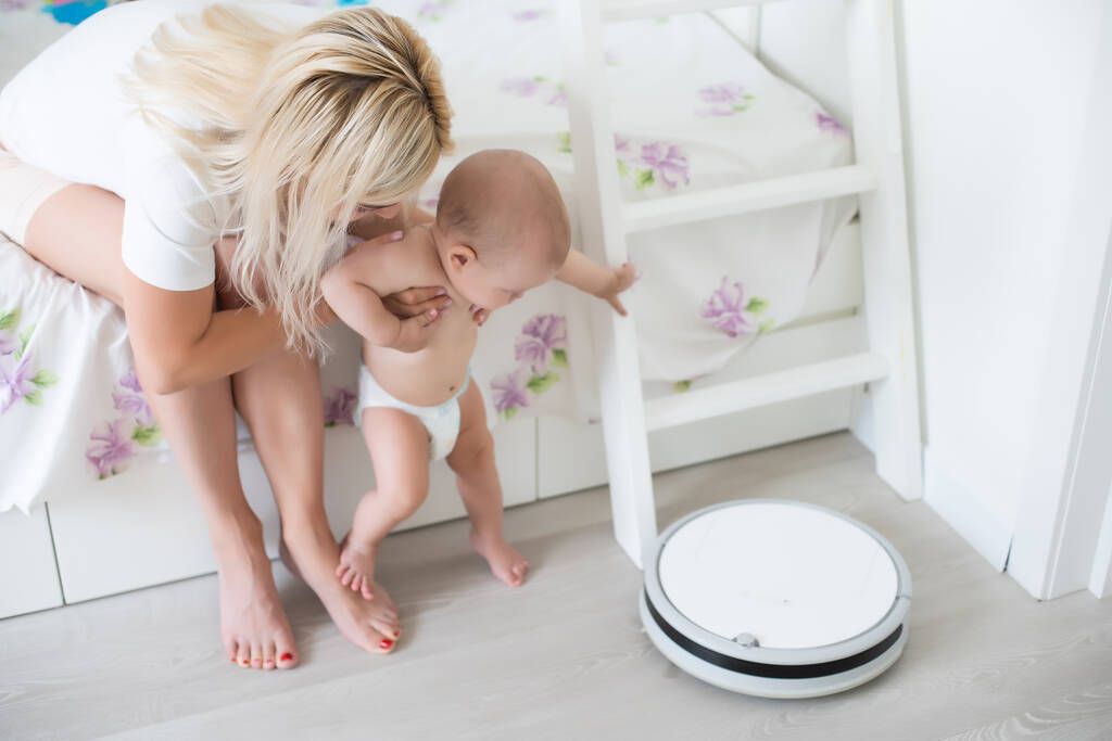 Женщина держит ребенка, а робот пылесос убирает комнату